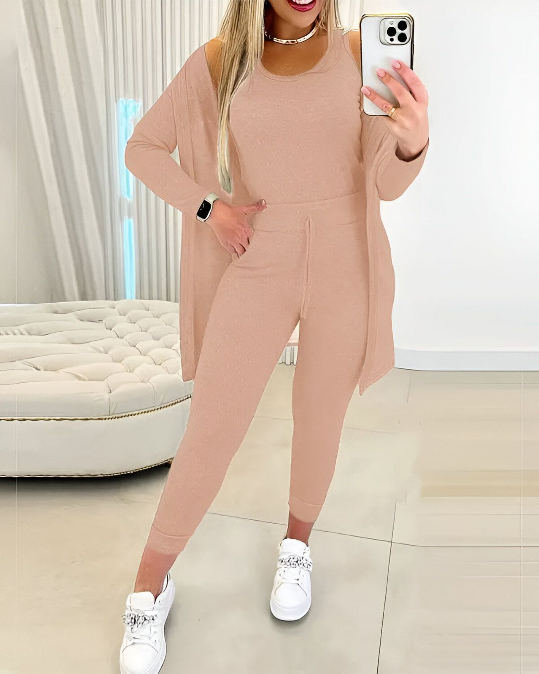 Une femme porte un ensemble beige composé d'un débardeur, d'un pantalon et d'un cardigan. Elle est debout dans une pièce et se prend en photo avec son téléphone. Elle porte des baskets blanches et un collier doré.