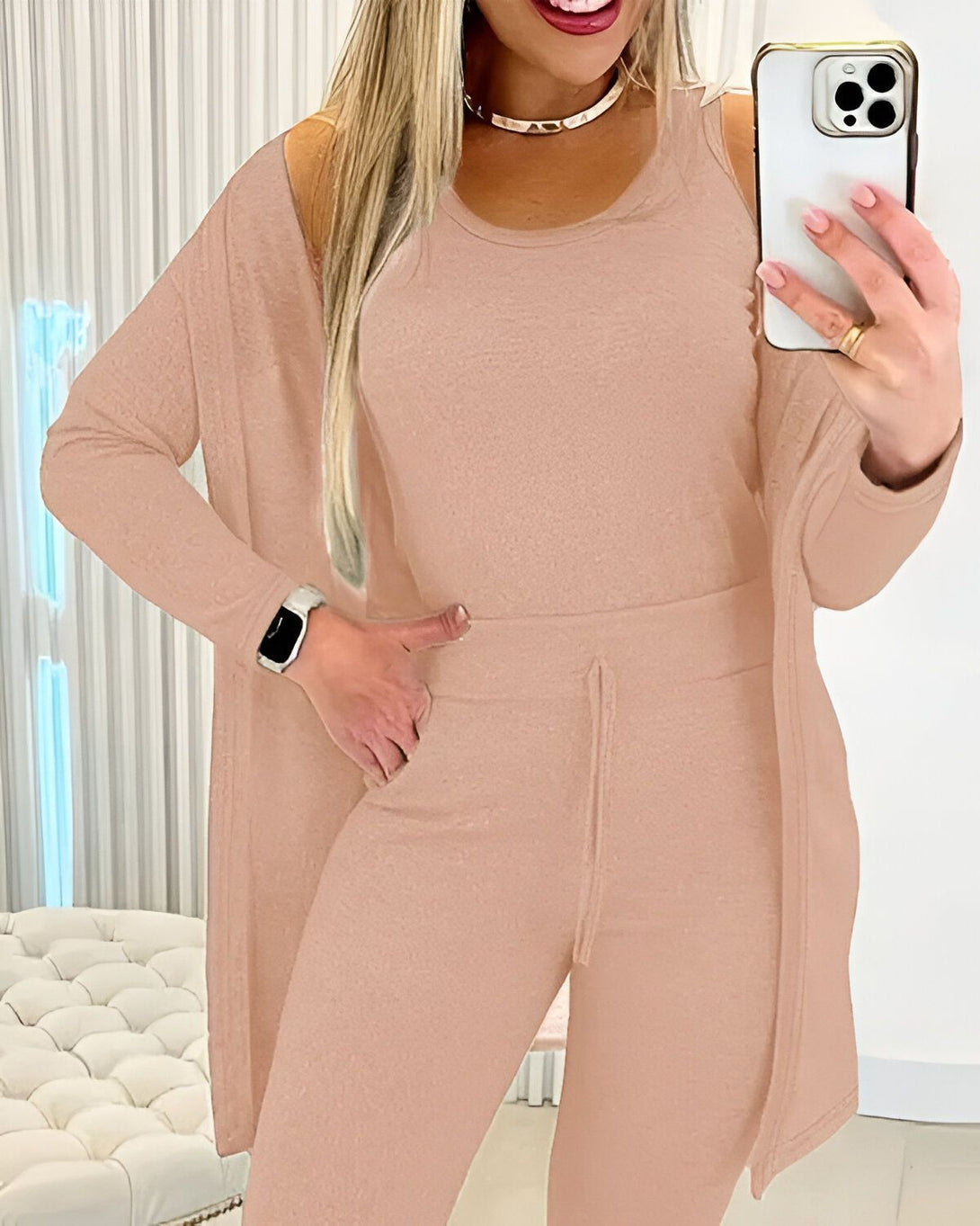 Une femme porte un ensemble beige composé d'un débardeur, d'un pantalon et d'un cardigan. Elle a une montre connectée au poignet gauche et tient un téléphone portable dans la main droite.