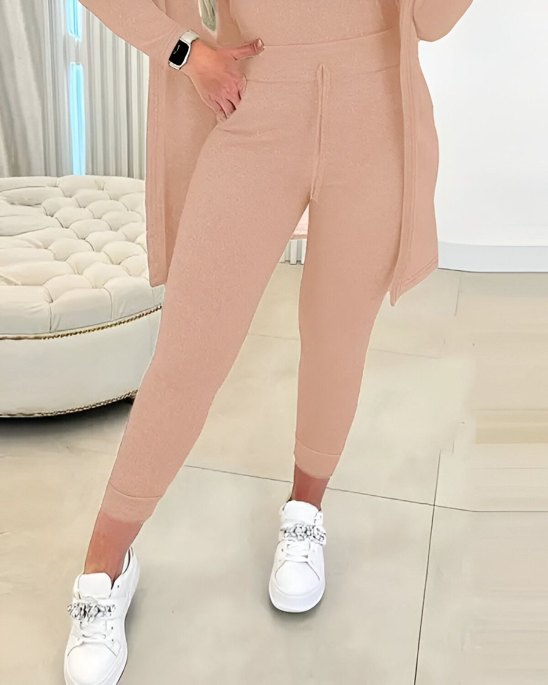 Une femme porte un ensemble jogging beige composé d'un pantalon de jogging beige et d'une veste beige ouverte. Elle porte également des baskets blanches.