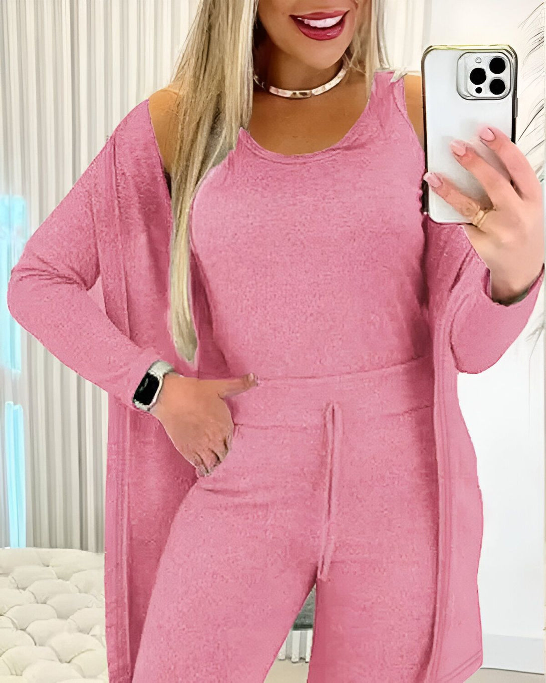 Une femme porte un ensemble trois pièces rose composé d'un débardeur, d'un pantalon et d'un cardigan. Elle a une montre connectée au poignet gauche et tient un téléphone portable dans sa main droite.