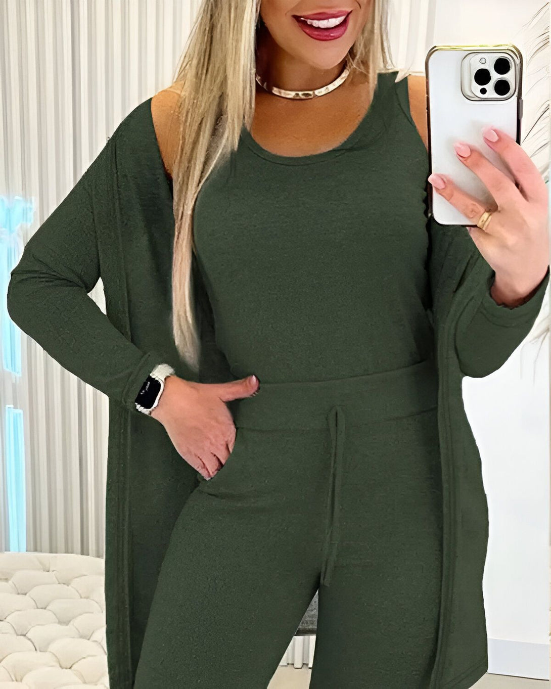 Une femme porte un ensemble trois pièces vert olive composé d'un débardeur, d'un pantalon et d'un cardigan. Elle a les cheveux blonds et porte un collier doré. Elle tient un téléphone portable dans sa main droite et a une montre connectée au poignet gauche.
