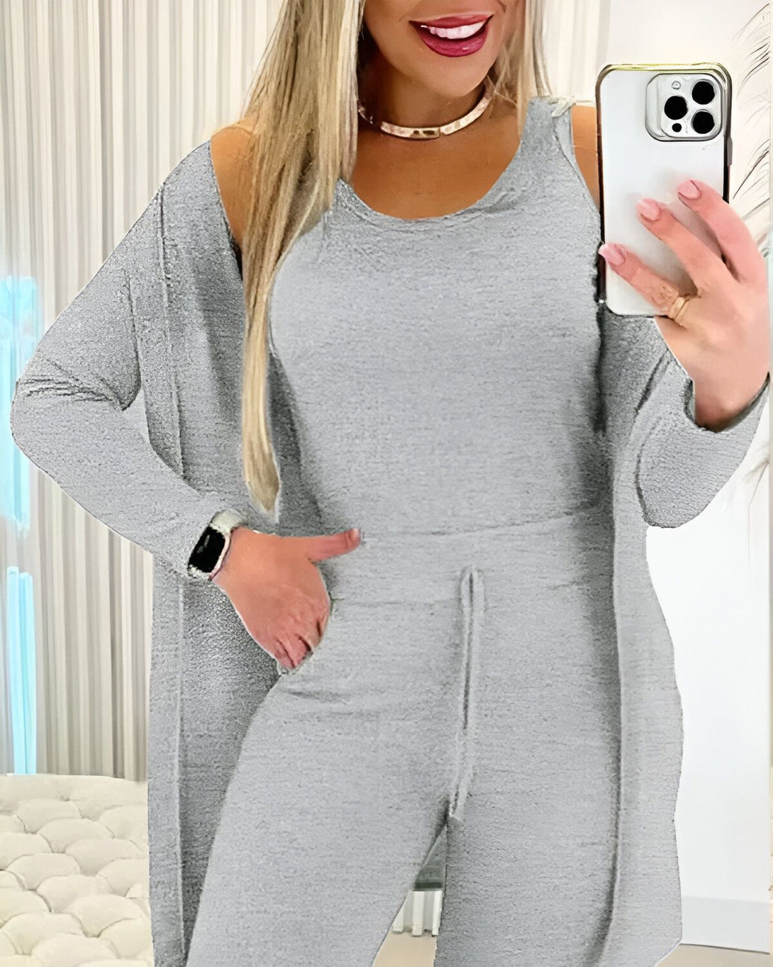 Une femme porte un ensemble jogging gris composé d'un débardeur, d'un pantalon et d'un cardigan. Elle a une montre connectée au poignet gauche et tient son téléphone portable dans la main droite.