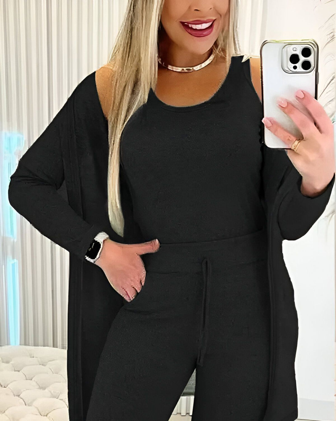 Une femme porte un ensemble trois pièces noir composé d'un débardeur, d'un pantalon et d'un cardigan. Elle est blonde, souriante et porte un collier doré. Elle tient un téléphone portable dans sa main droite et se prend en photo dans un miroir.