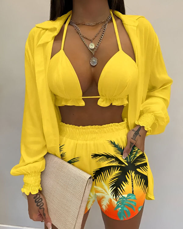 Une femme porte un ensemble jaune composé d'un short, d'un soutien-gorge et d'une chemise ouverte. Le short est imprimé de feuilles de palmier vertes et le soutien-gorge est jaune uni. La chemise est ouverte et nouée au niveau de la taille. La femme porte également un collier et un sac à main jaunes.