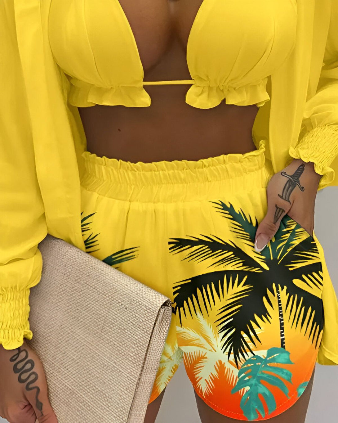Une femme porte un ensemble deux pièces jaune composé d'un crop top et d'un short. Le crop top est en mousseline de soie jaune et possède des manches longues et bouffantes. Le short est taille haute et possède une ceinture élastique. Il est imprimé de feuilles de palmier tropicales colorées. La femme porte également un sac à main beige et des talons hauts jaunes.