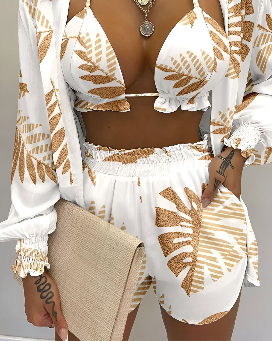 Une femme porte un ensemble trois pièces blanc et marron. Le haut est un soutien-gorge triangle, le short est taille haute et la veste est ouverte. L'ensemble est imprimé de feuilles tropicales.