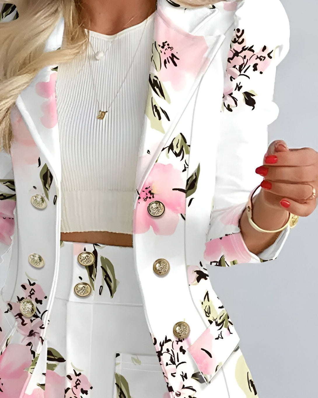 Une femme porte un tailleur blanc à motifs floraux roses. Le tailleur est composé d'une veste et d'un pantalon. La veste est cintrée et possède des boutons dorés. Le pantalon est droit et possède des poches. La femme porte également un collier et des bracelets.