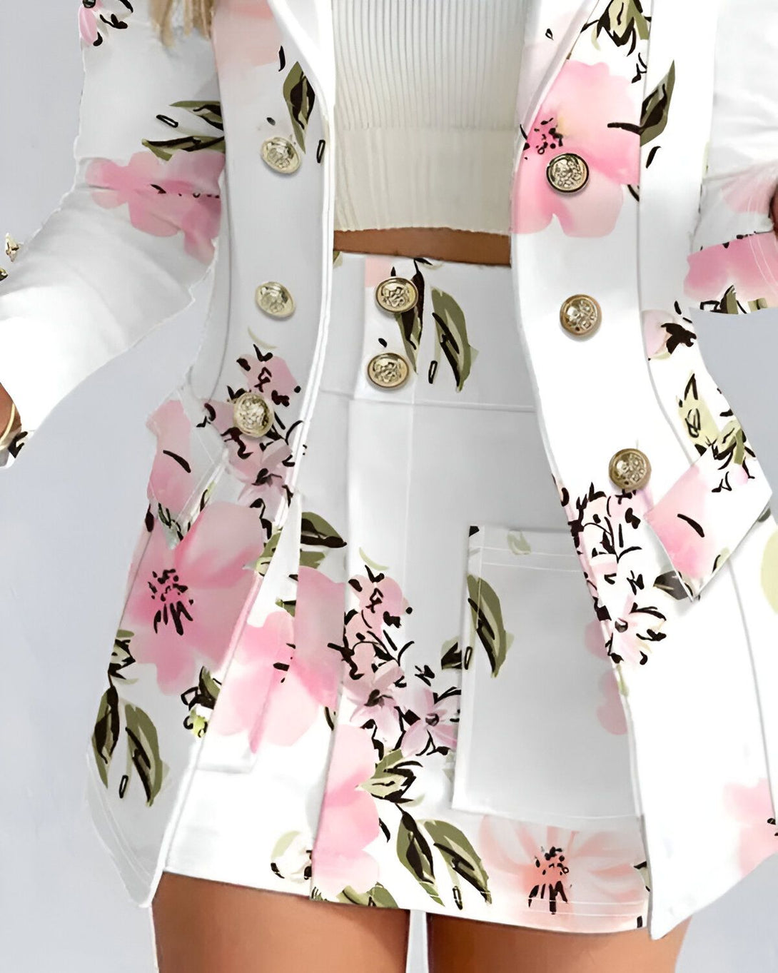 Une femme porte un tailleur blanc à motifs floraux roses. Le tailleur est composé d'une veste et d'une jupe. La veste est cintrée et possède des boutons dorés. La jupe est droite et possède des poches.