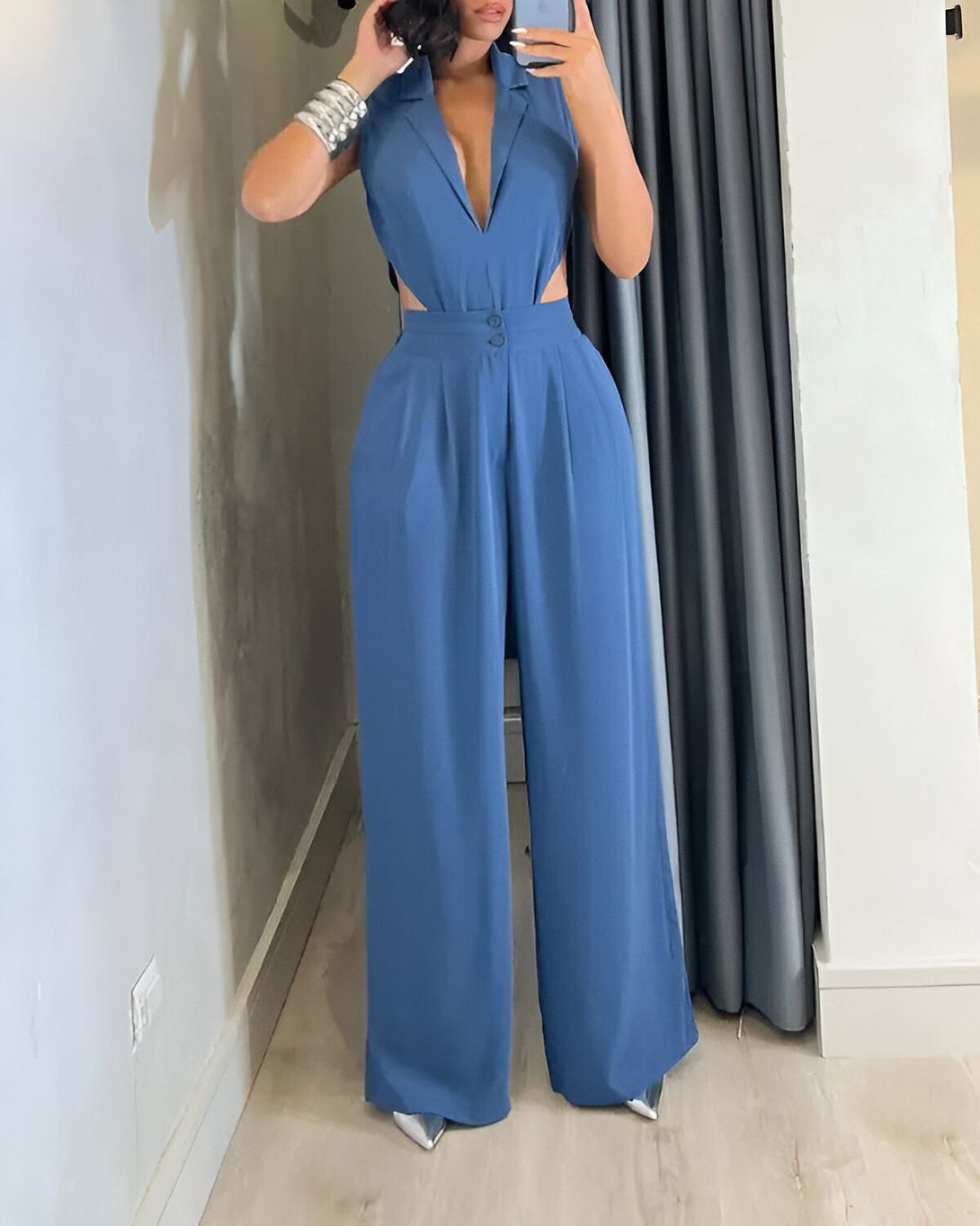 Une femme porte une combinaison pantalon bleu ciel. La combinaison a un col en V et des jambes larges. Elle est cintrée à la taille et possède des boutons sur le devant. La femme porte des escarpins argentés et des bracelets aux poignets.