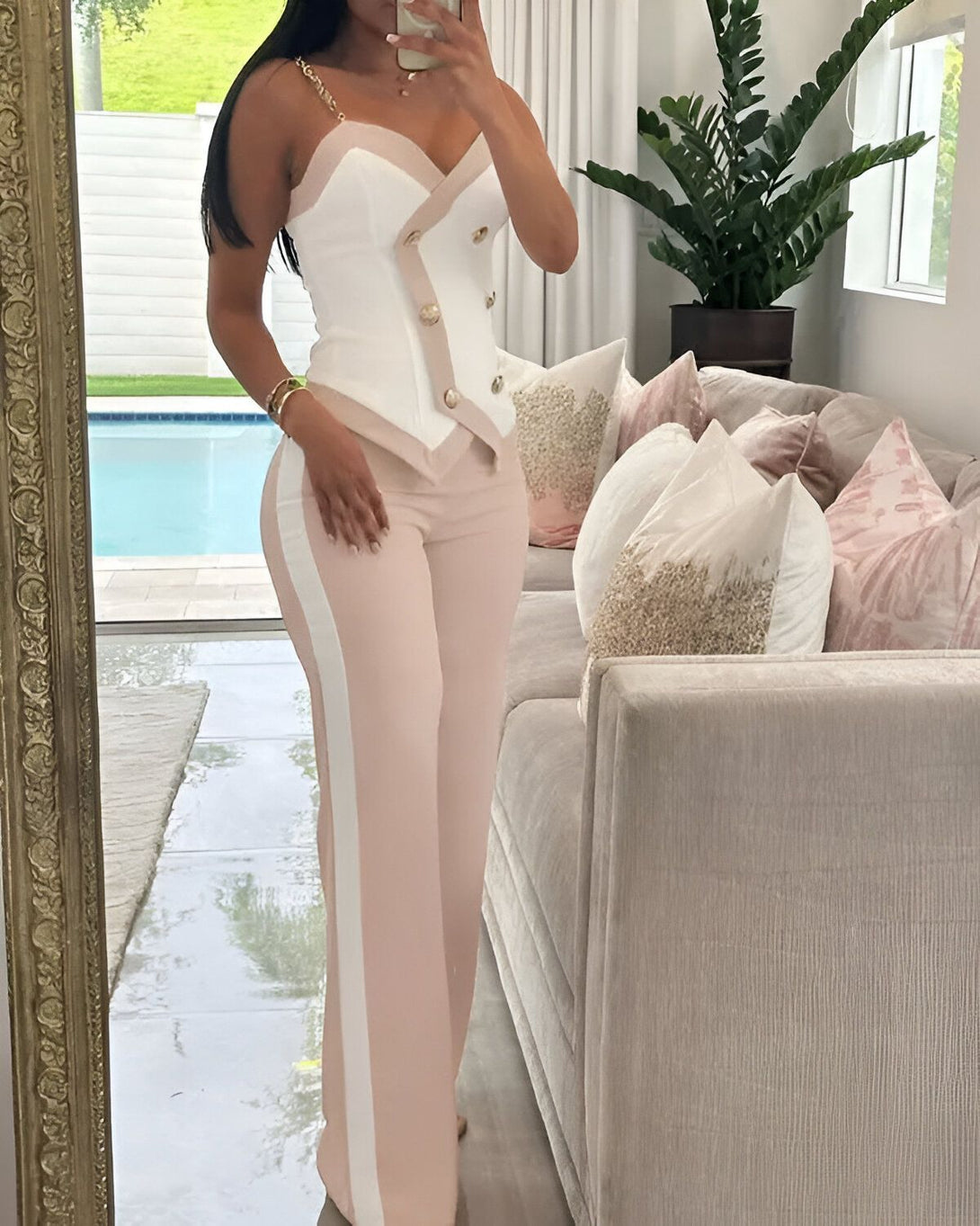 Une femme porte un tailleur pantalon beige et blanc. Le haut est un bustier avec des boutons dorés. Le pantalon est taille haute et évasé. La femme est debout dans un salon, il y a un canapé et une plante verte derrière elle.