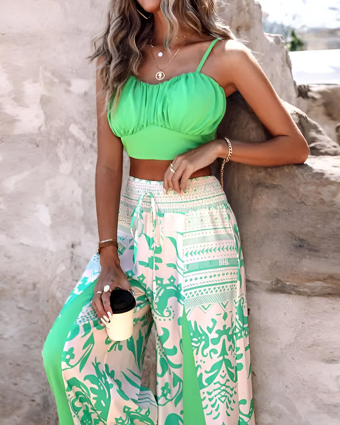 Une femme porte un haut vert et un pantalon large à motifs. Elle a les cheveux longs et blonds et porte un collier. Elle tient une tasse à café à la main.