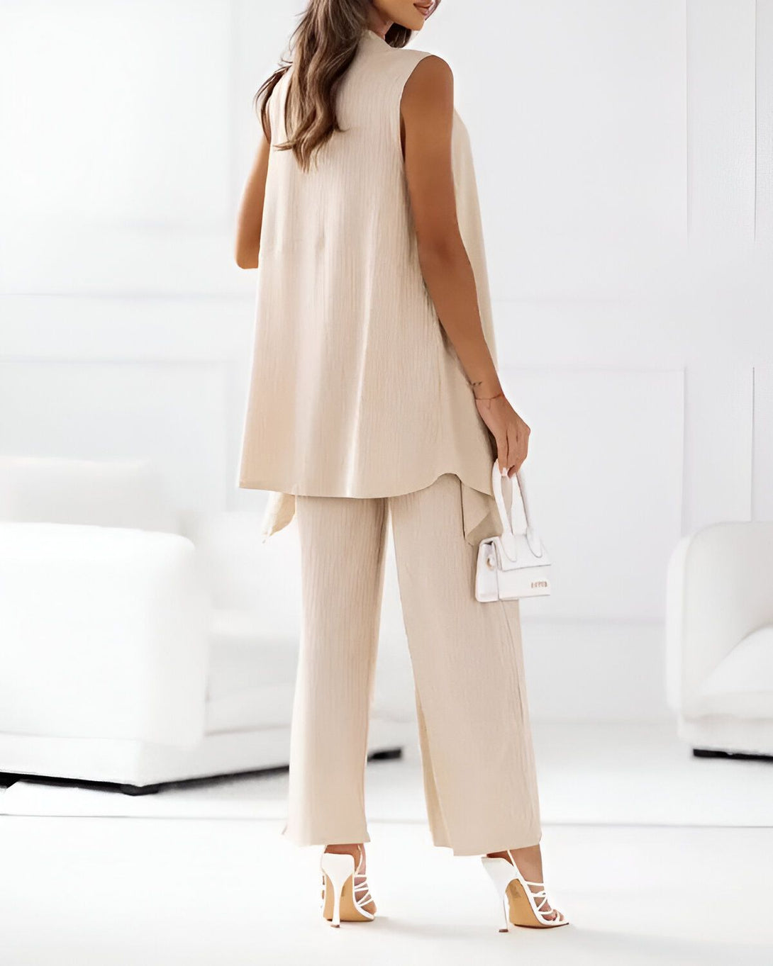 Une femme porte un ensemble pantalon et gilet beige et des sandales blanches à talons. Elle porte également un sac à main blanc.