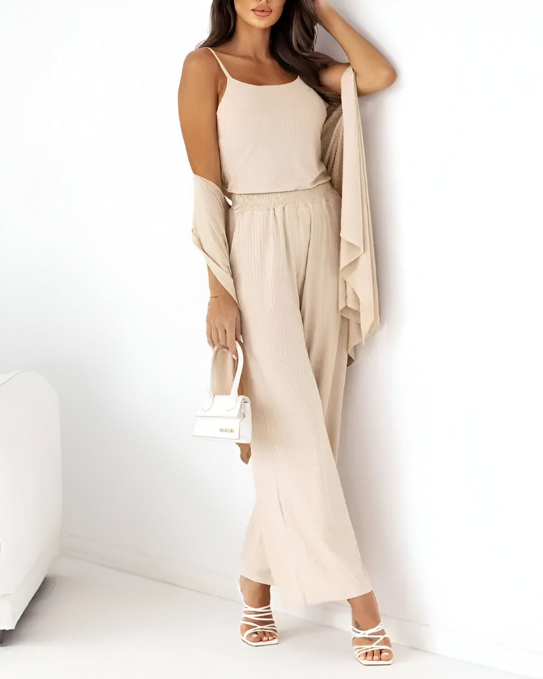 Une femme porte un pantalon beige large et un débardeur beige. Elle porte également un cardigan beige et des sandales à talons blancs. Elle porte un sac à main blanc à la main.