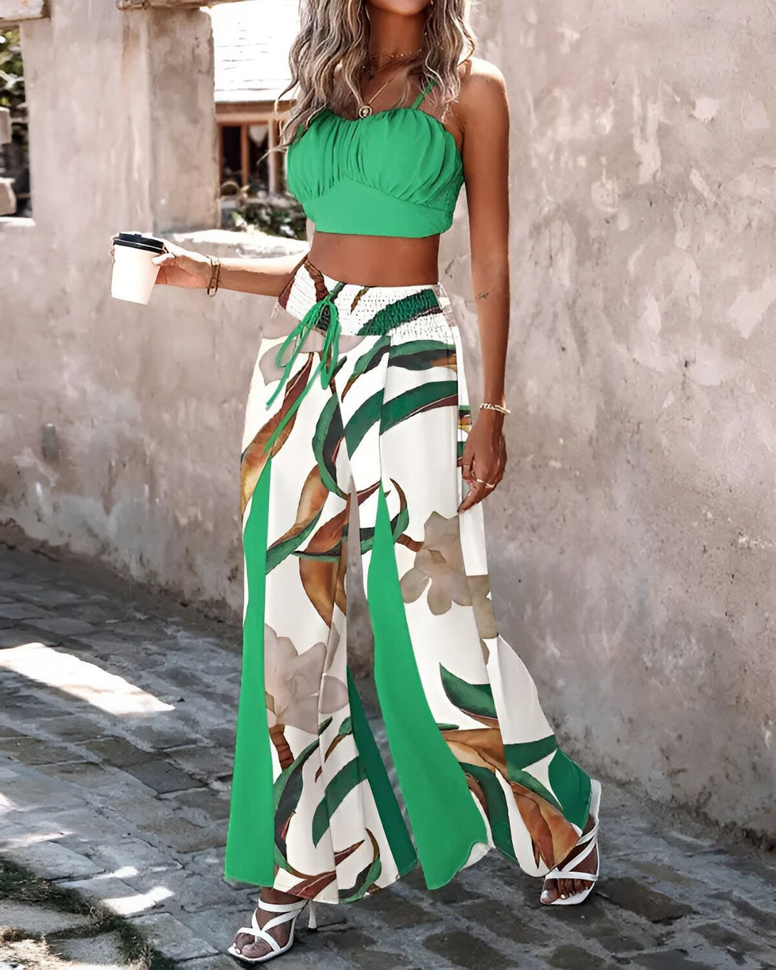 Une femme porte un haut court vert et un pantalon large imprimé de feuilles tropicales. Elle porte également des sandales à talons hauts et tient une tasse de café à la main.