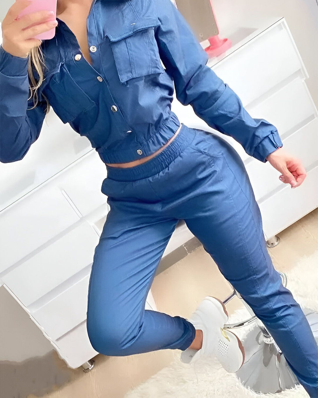 Une femme porte un ensemble de jogging bleu composé d'une veste et d'un pantalon. La veste est courte et possède des boutons. Le pantalon est taille haute et possède des poches. La femme est debout et prend une photo d'elle-même dans un miroir.