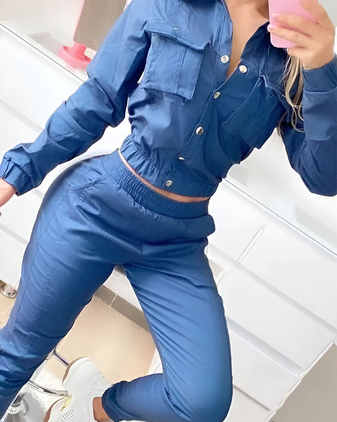 Une femme porte un ensemble de jogging bleu marine. L'ensemble est composé d'une veste et d'un pantalon. La veste est courte et possède des boutons. Le pantalon est taille haute et possède des poches. La femme est debout et prend une photo d'elle-même dans un miroir.