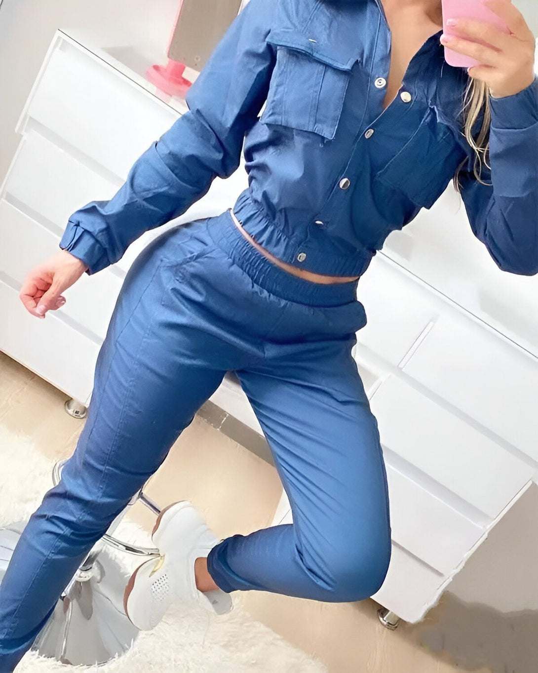 Une femme porte un ensemble de jogging bleu composé d'une veste et d'un pantalon. La veste est ouverte et on peut voir un haut bleu marine en dessous. Elle porte des baskets blanches et se prend en photo dans un miroir.