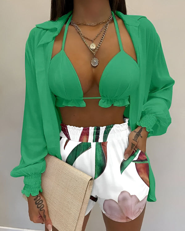Une femme porte un haut de bikini vert, un short blanc à motifs tropicaux et une chemise verte transparente.