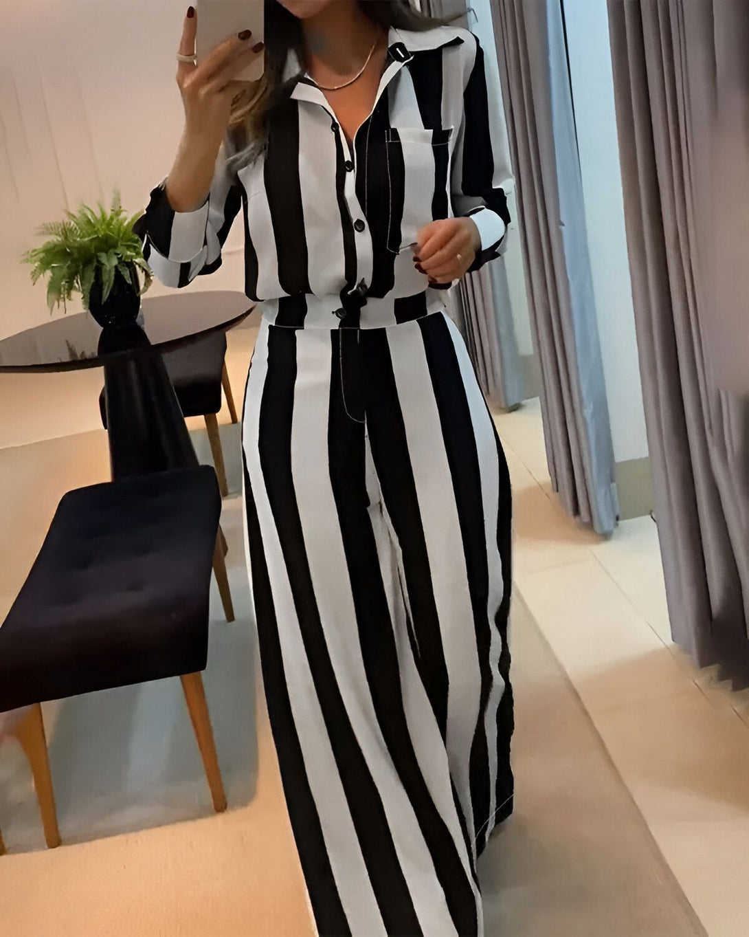 Une femme porte un chemisier et un pantalon rayés noirs et blancs. Elle est debout dans une pièce, il y a une plante verte sur une table à sa gauche et des rideaux blancs à sa droite.