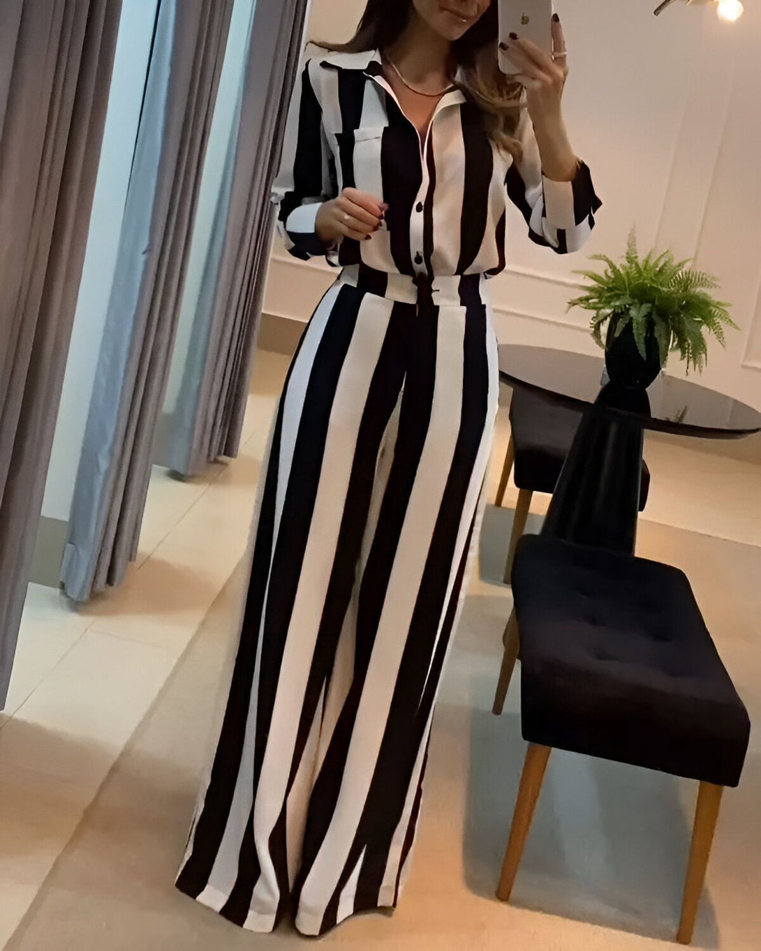 Une femme porte un chemisier et un pantalon rayés noirs et blancs. Elle est debout dans une pièce, il y a une plante verte et une chaise noire derrière elle.