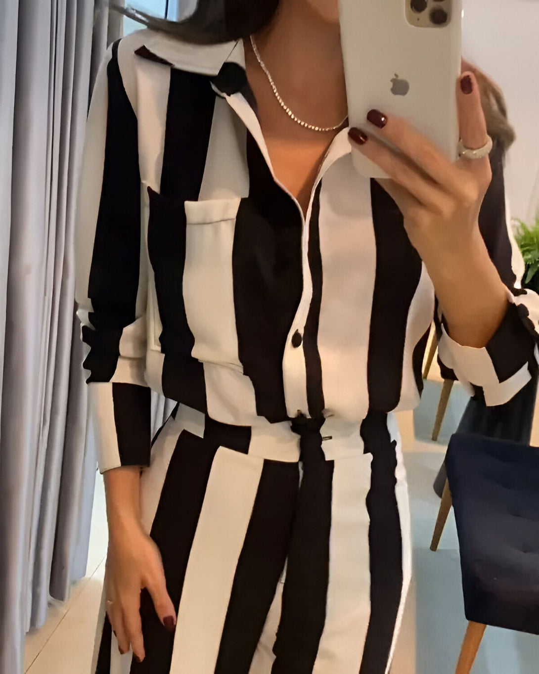 Une femme porte un tailleur pantalon noir et blanc à rayures verticales. Elle a une chemise ouverte avec un col et des manches longues, et un pantalon assorti avec une ceinture. Elle porte également un collier et des boucles d'oreilles.