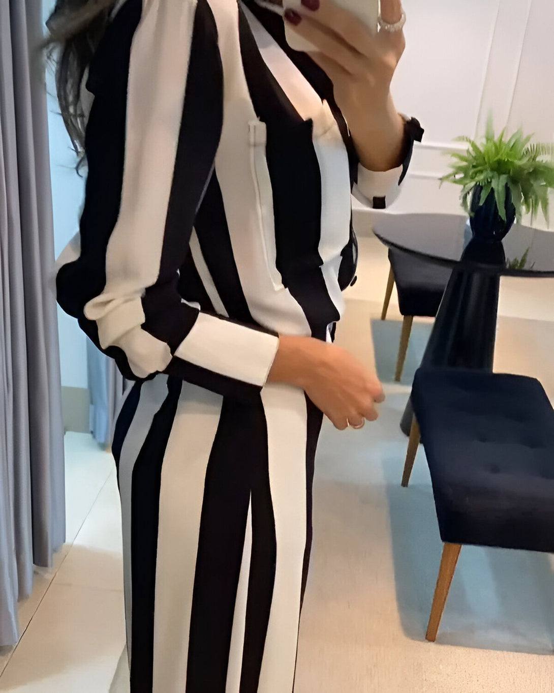 Une femme porte un tailleur pantalon noir et blanc à rayures verticales. Elle est vue de dos et tient son téléphone portable à la main. A côté d'elle se trouve une chaise noire et une plante verte.