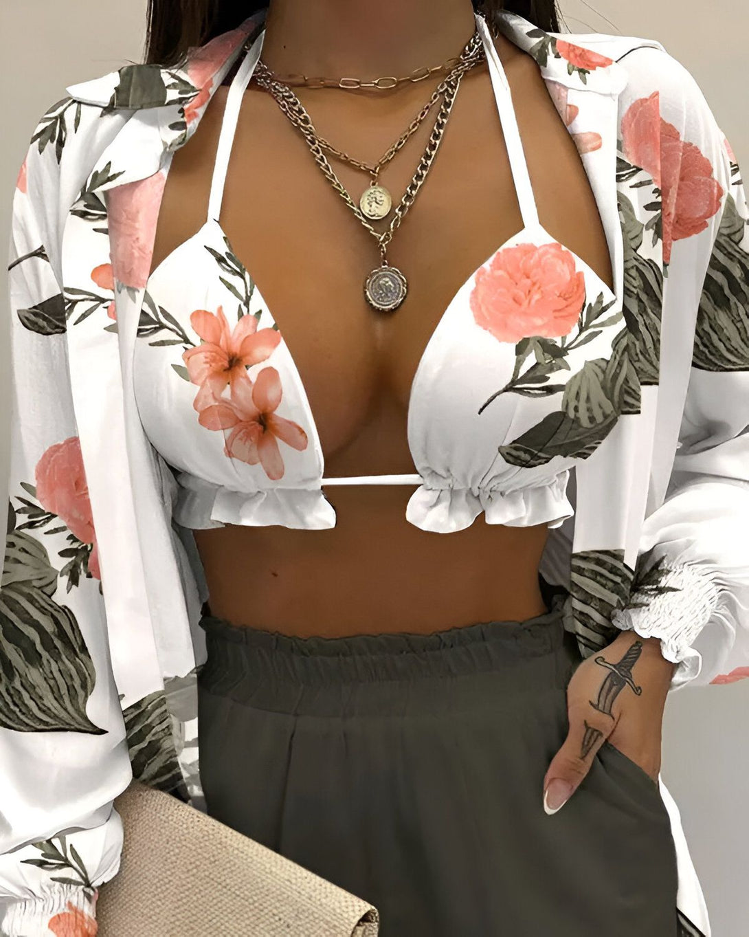 Une femme porte un haut de bikini triangle à fleurs avec un short taille haute kaki et une chemise ouverte blanche à manches longues. Elle porte également plusieurs colliers et un sac à main beige.