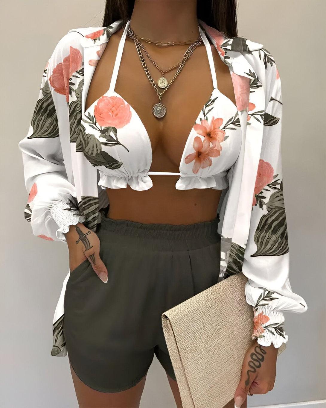 Une femme porte un haut de bikini blanc à fleurs roses, un short vert kaki et une chemise blanche à fleurs roses. Elle porte également un collier avec un pendentif rond et une chaîne en or. Elle a un sac à main beige dans la main droite.