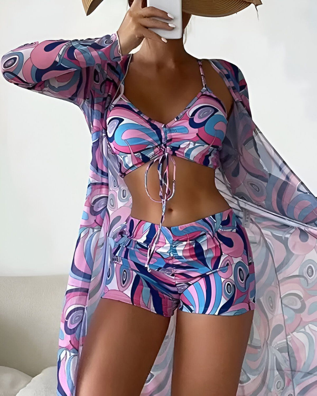 Une femme porte un maillot de bain trois pièces coloré avec un motif psychédélique. Le haut est un soutien-gorge triangle avec des bonnets froncés et des bretelles fines. Le bas est un short taille haute avec des liens à nouer sur les côtés. La femme porte également une longue robe de plage transparente par-dessus son maillot de bain. Elle a un chapeau de paille sur la tête et tient un téléphone portable à la main.