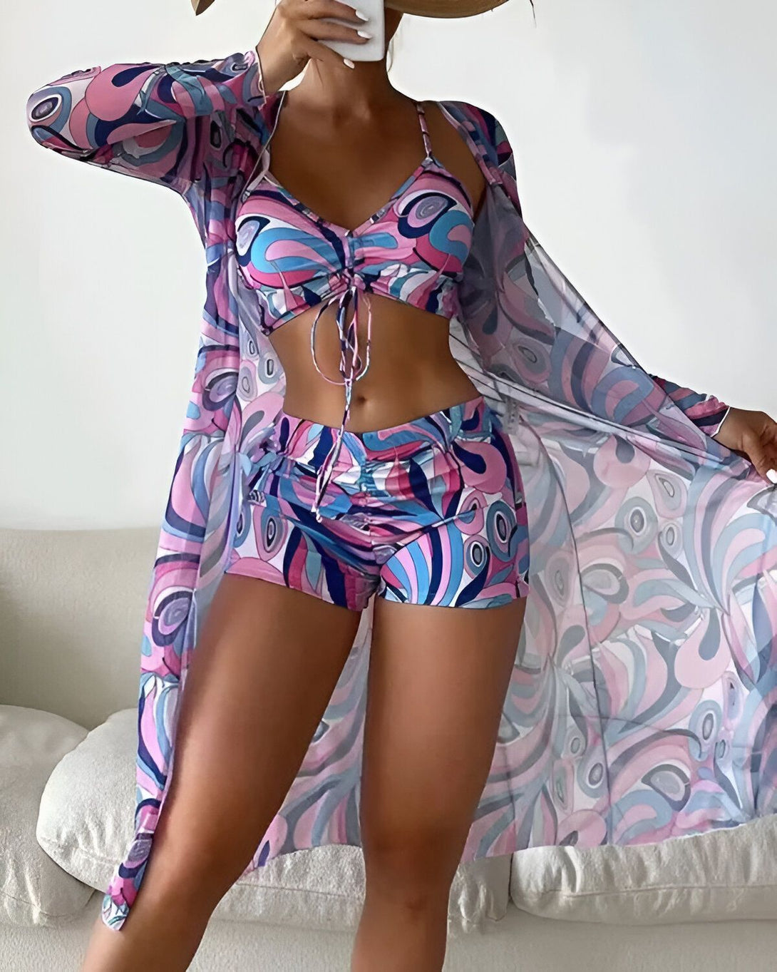 Une femme porte un maillot de bain trois pièces coloré avec un motif psychédélique. Le haut est un soutien-gorge triangle avec des bonnets froncés et des bretelles fines. Le bas est un short taille haute avec des liens à nouer sur les côtés. La femme porte également une longue robe de plage transparente par-dessus son maillot de bain.