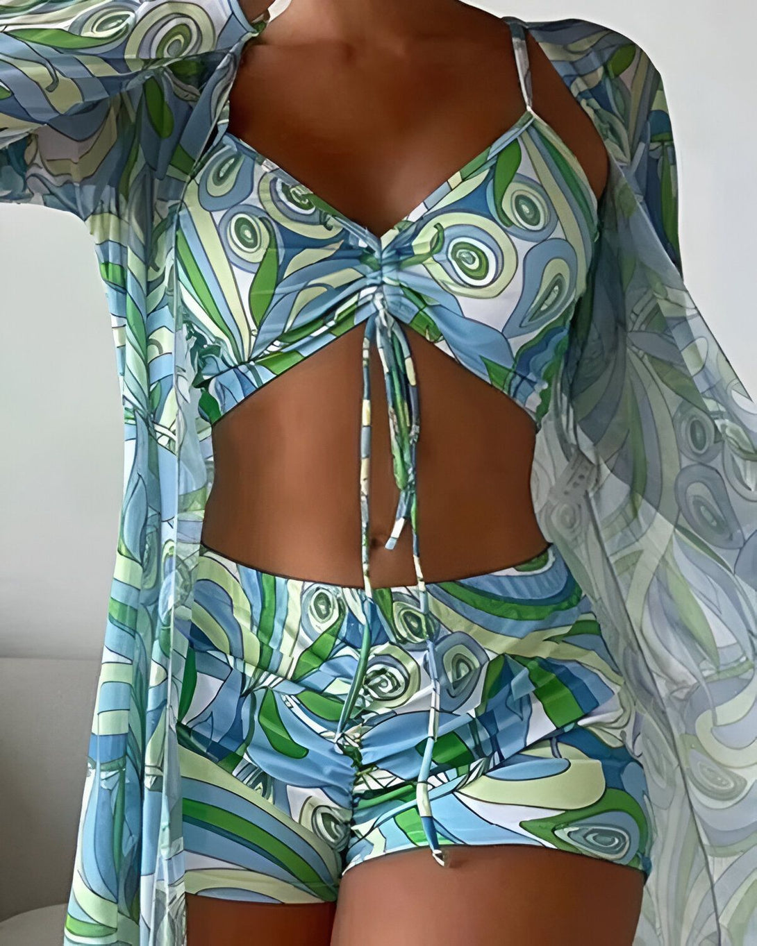 Une femme porte un ensemble de plage trois pièces bleu et vert. Il est composé d'un short, d'un haut et d'une veste ouverte. Le short et le haut sont de la même couleur et ont un motif de cercles concentriques. La veste est ouverte et a le même motif que le short et le haut.