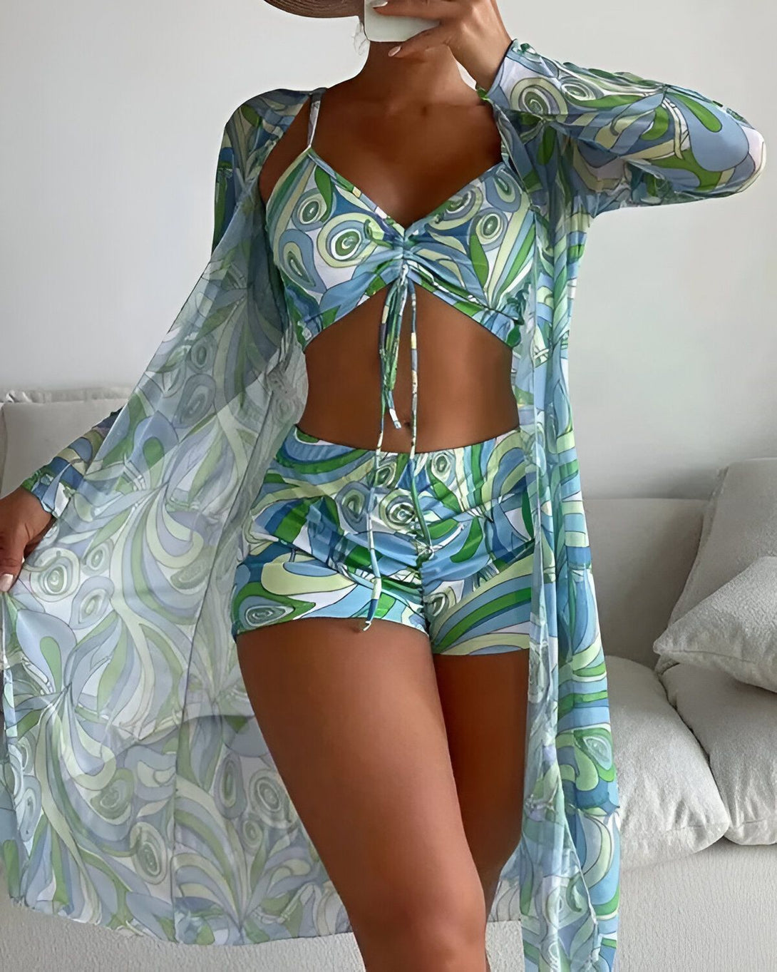 Une femme porte un ensemble de plage trois pièces bleu et vert. Il est composé d'un short, d'un haut et d'un kimono. Le short et le haut sont de la même couleur et ont un motif psychédélique. Le kimono est bleu transparent et a un motif de vagues.