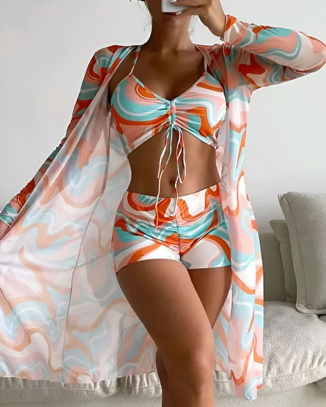 Une femme porte un ensemble de plage trois pièces de couleur orange et bleu. Il est composé d'un short, d'un haut et d'un kimono. Le short est taille haute et possède une coupe évasée. Le haut est un crop top à nouer au niveau de la poitrine. Le kimono est long et transparent.