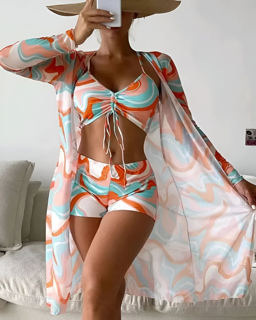Une femme porte un maillot de bain trois pièces avec un motif coloré. Le haut est un soutien-gorge triangle avec des bonnets froncés et des bretelles fines. Le bas est un shorty taille haute avec des liens à nouer sur les côtés. Le paréo est long et transparent, avec des manches longues et un motif coloré assorti au maillot de bain.