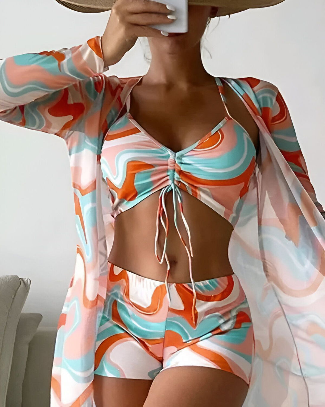 Une femme porte un ensemble de plage trois pièces coloré. Le haut est un crop top à motif cache-cœur avec des bonnets froncés et des bretelles fines. Le short est taille haute et possède une coupe évasée. La veste est longue et ouverte, avec des manches longues et fluides. L'ensemble est de couleur orange, rose et bleu.