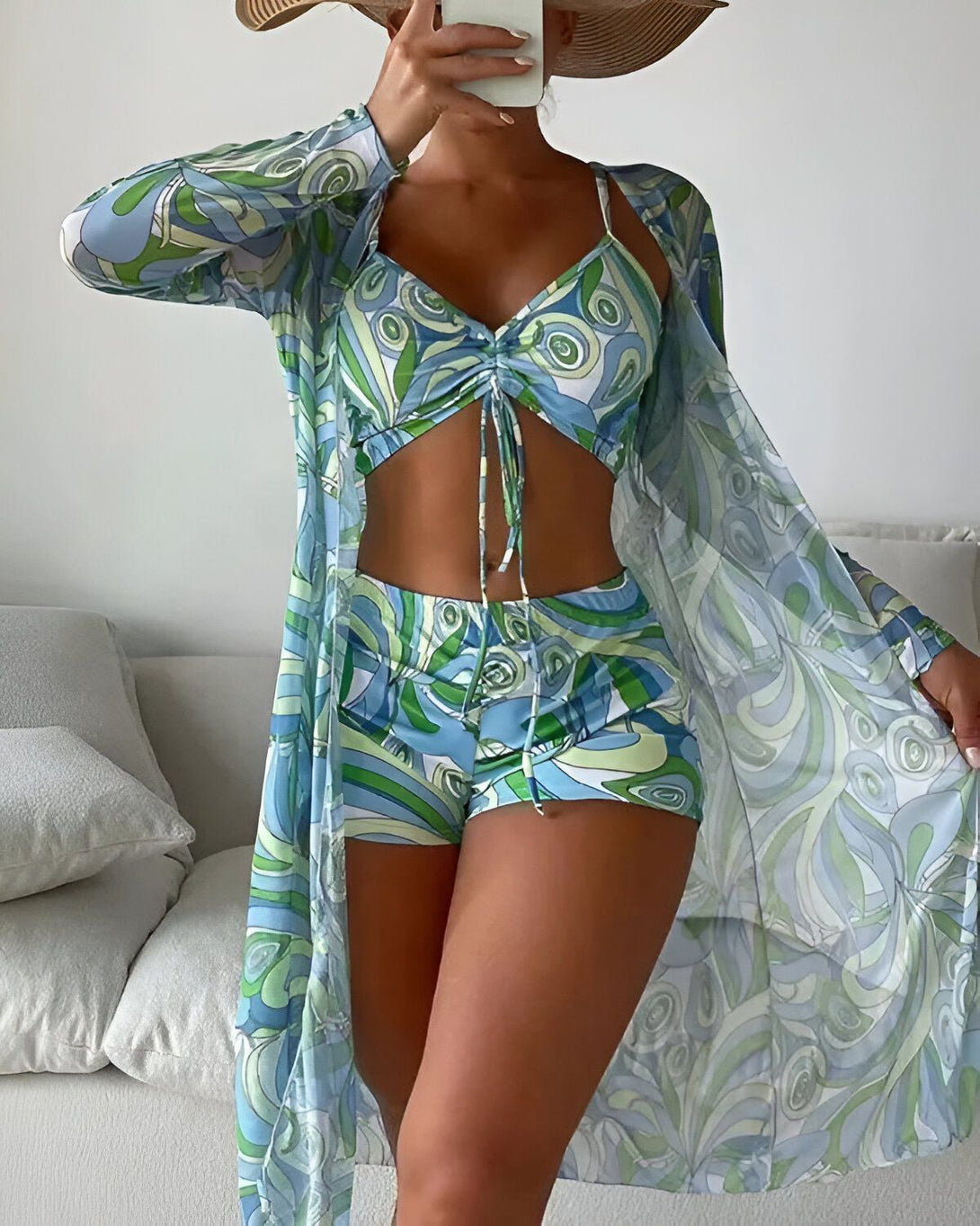 Une femme porte un maillot de bain trois pièces bleu et vert. Le haut est un soutien-gorge triangle à armatures avec des bonnets froncés. Le bas est un shorty taille haute. Le paréo est long et ample, avec des manches kimono. La femme est debout devant un miroir et se prend en photo.