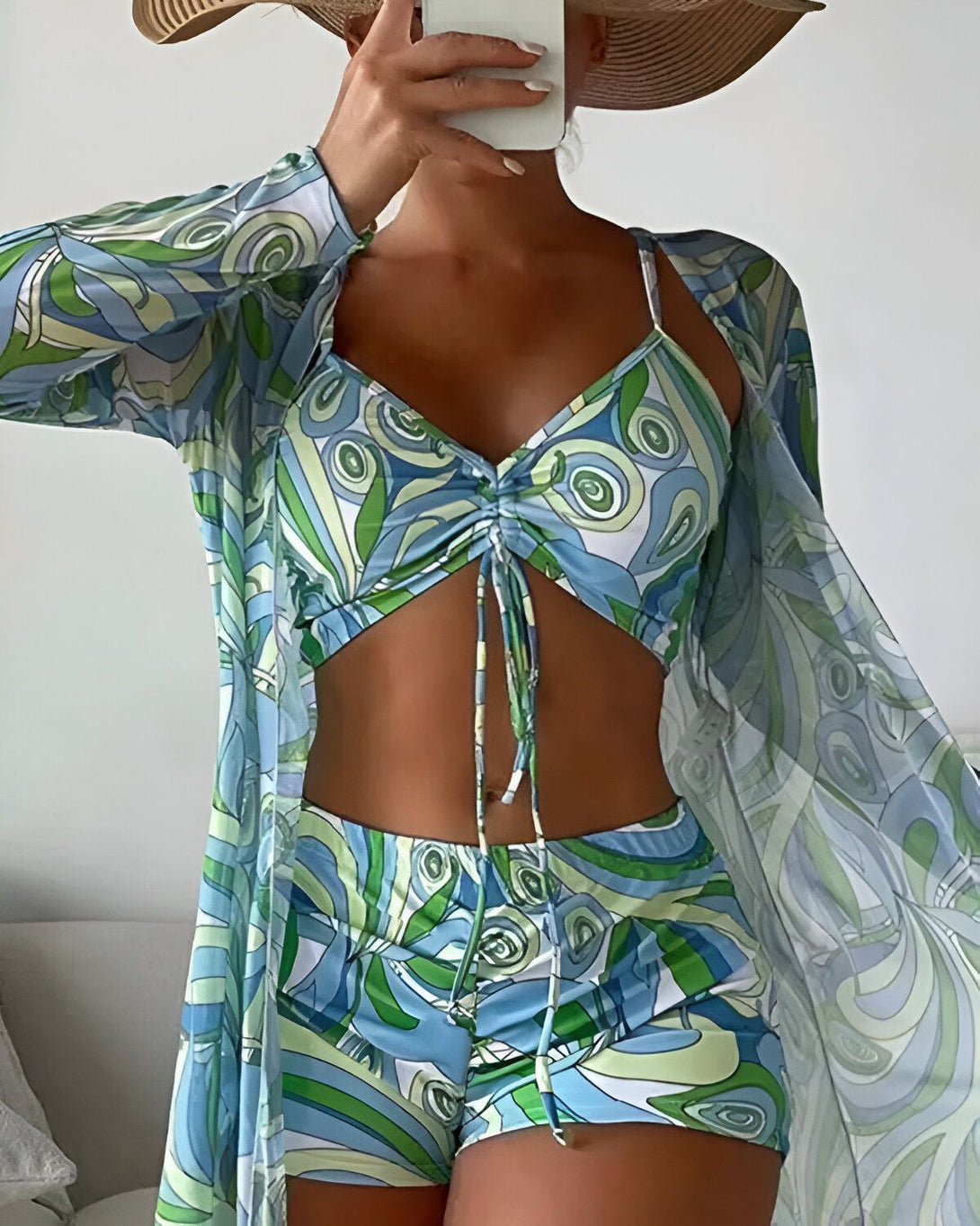 Une femme porte un ensemble de plage trois pièces bleu et vert. Il est composé d'un short, d'un haut et d'une veste ouverte. Le haut est un crop top à bretelles fines. Le short est taille haute et échancré. La veste est longue et ample.
