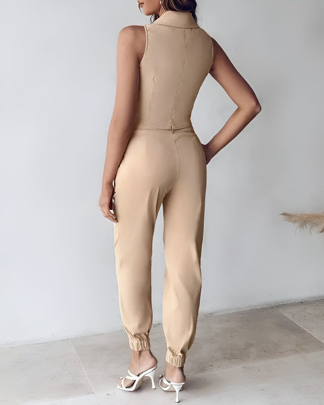 Une femme porte un ensemble pantalon et haut beige. Le haut est court et sans manches, avec un col montant. Le pantalon est ample et possède des poches latérales. La femme est vue de dos.