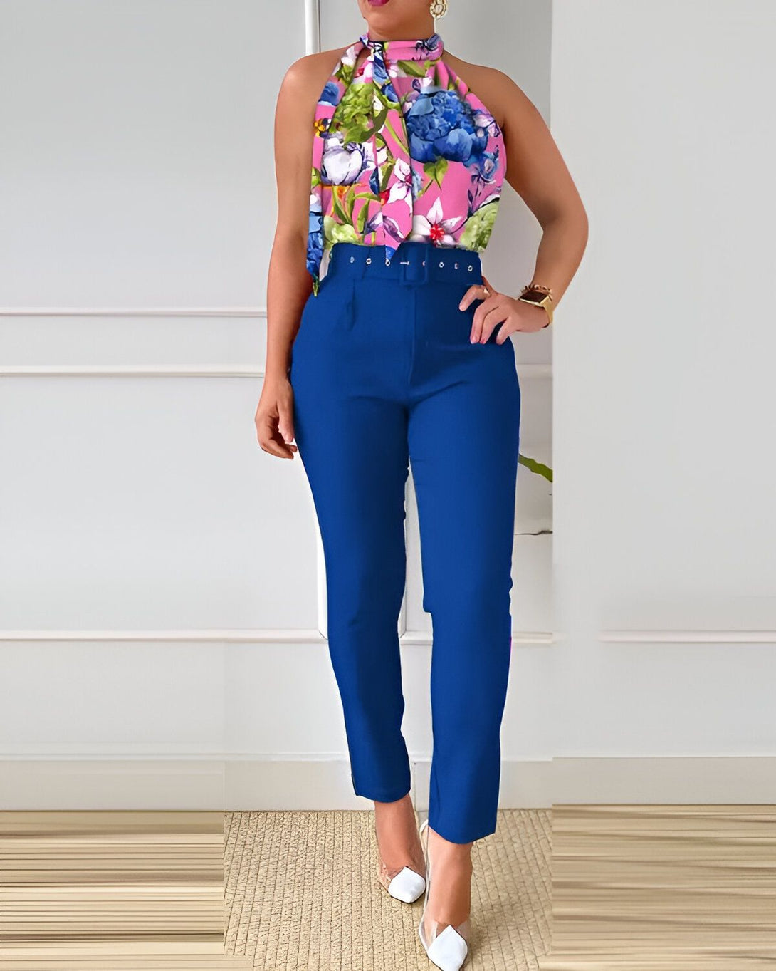 Une femme porte un pantalon bleu et un haut à fleurs rose et bleu. Elle est debout dans une pièce blanche.