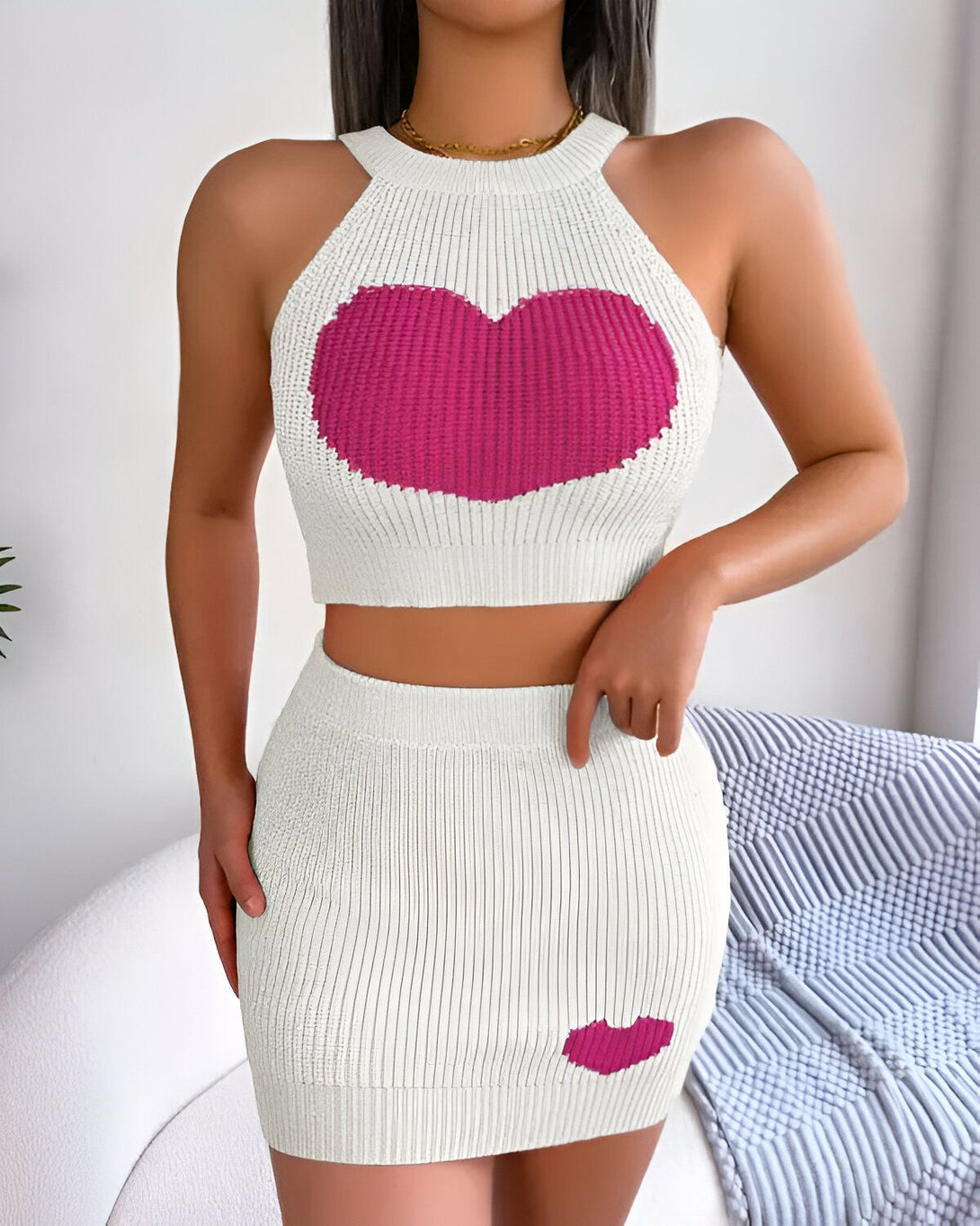 Une femme porte un ensemble de deux pièces en tricot blanc avec un motif de cœur rose. Le haut est un crop top avec une encolure licou et le bas est une jupe courte.