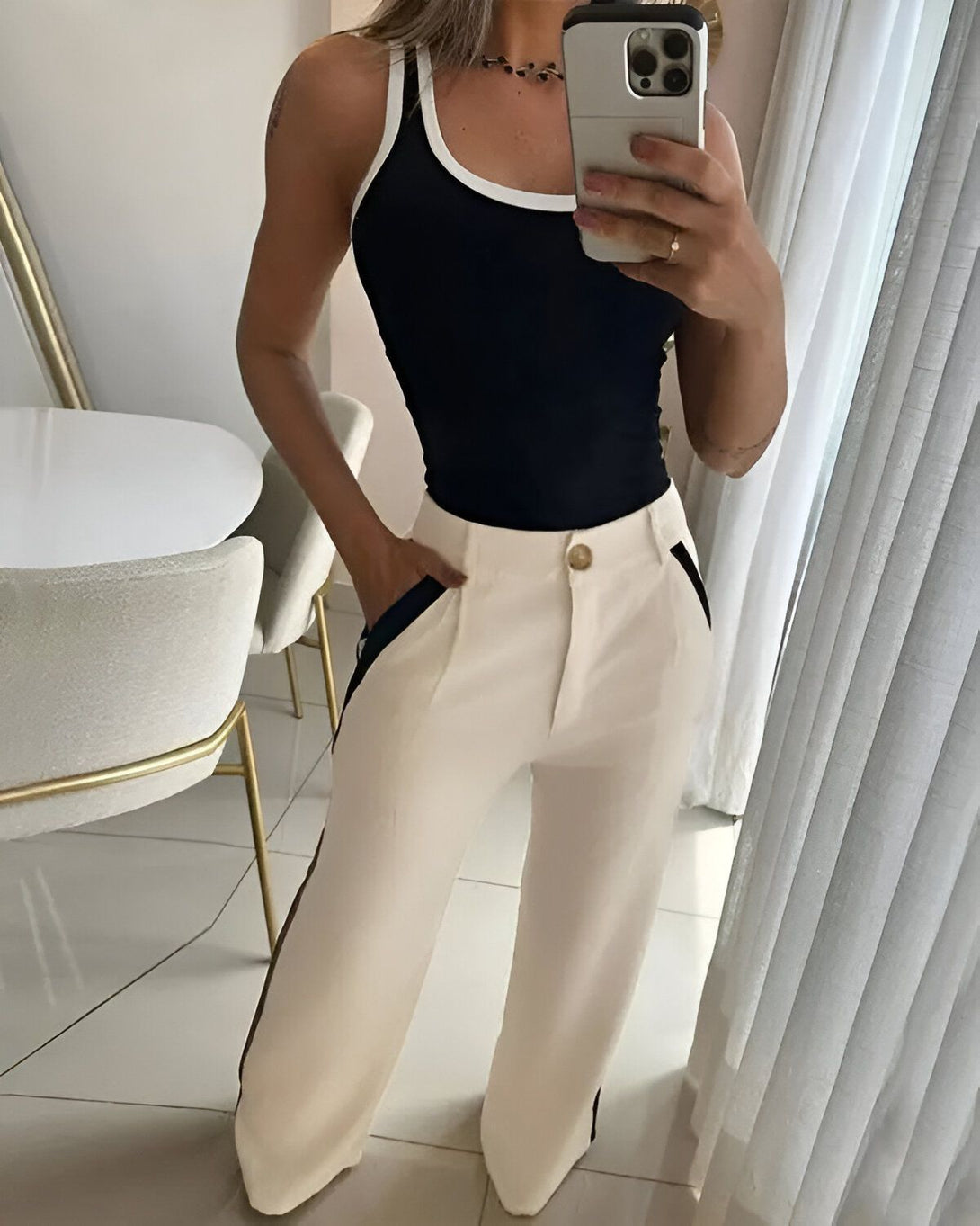 Une femme porte un débardeur noir et un pantalon blanc. Elle est debout dans une pièce, et tient son téléphone dans sa main droite.