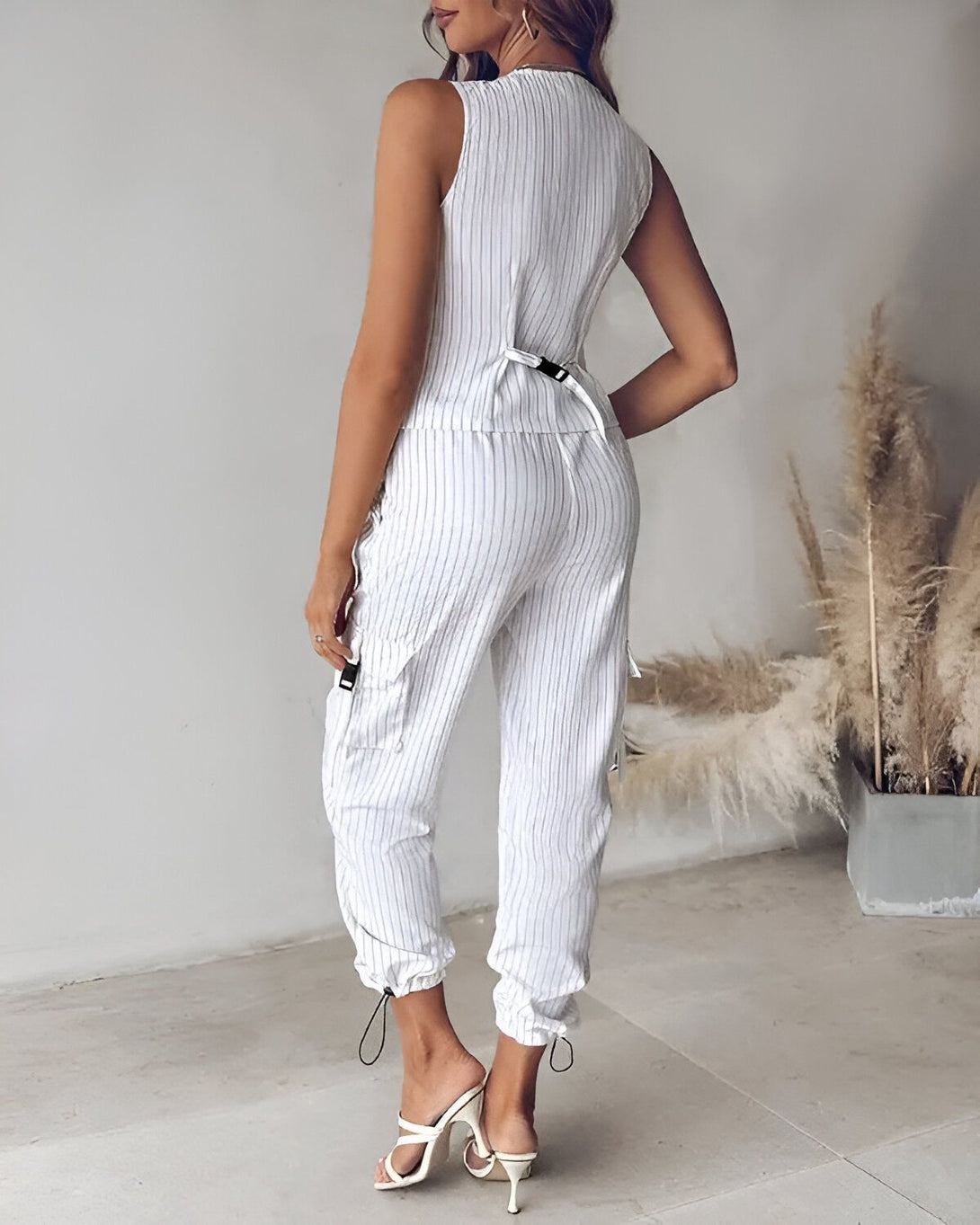 Une femme porte un pantalon cargo blanc à rayures verticales et un débardeur blanc. Elle est vue de dos.