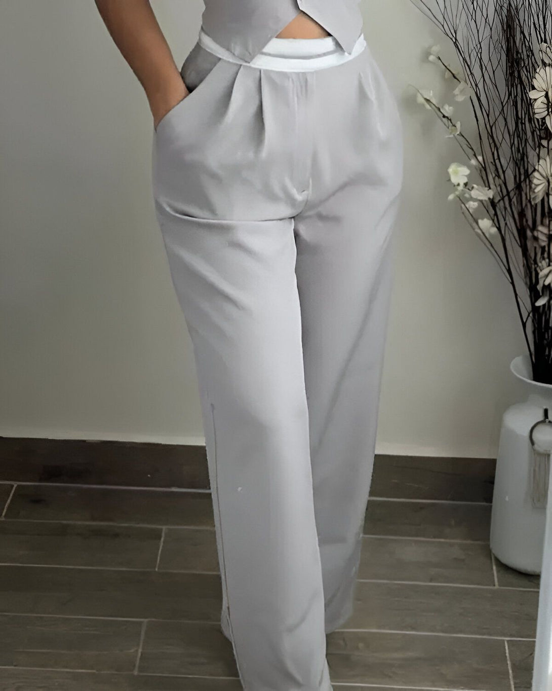 Une femme porte un pantalon gris taille haute avec une ceinture blanche. Le pantalon est ample et a des plis sur le devant. La femme est debout dans une pièce avec un mur blanc en arrière-plan.