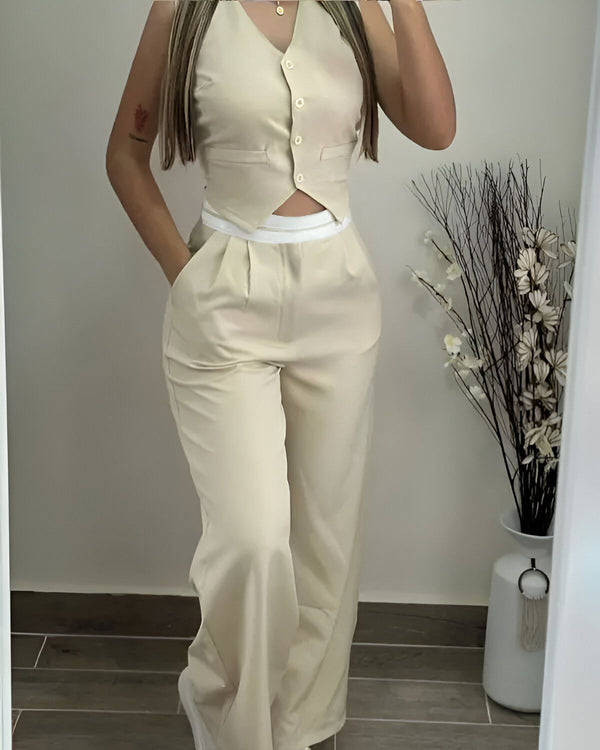 Une femme porte un gilet beige et un pantalon beige. Le gilet est court et ajusté, et le pantalon est ample et taille haute. La femme est debout dans un salon, et il y a un vase avec des fleurs blanches en arrière-plan.