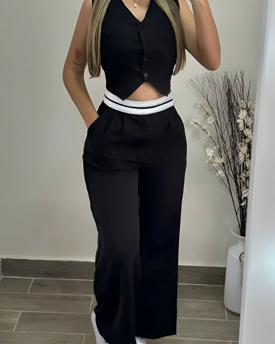 Une femme porte un gilet noir et un pantalon noir. Le gilet est court et a des boutons blancs. Le pantalon est large et a une taille haute. La femme est debout dans un miroir et se prend en photo.