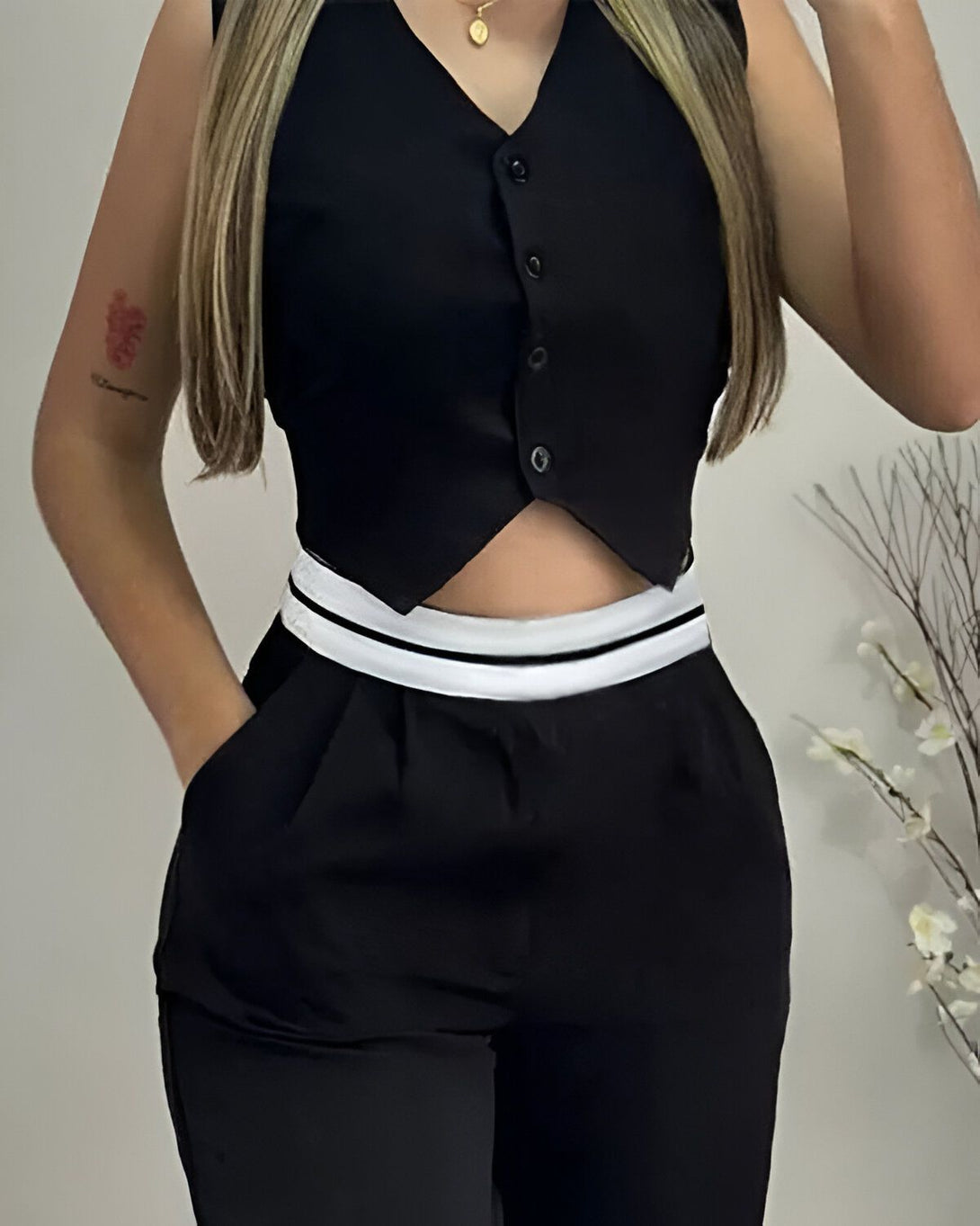 Une femme porte un gilet noir et un pantalon noir. Le gilet est court et cintré, avec des boutons blancs. Le pantalon est taille haute et a une coupe droite. La femme est debout, les mains dans les poches. Elle a les cheveux longs et blonds.