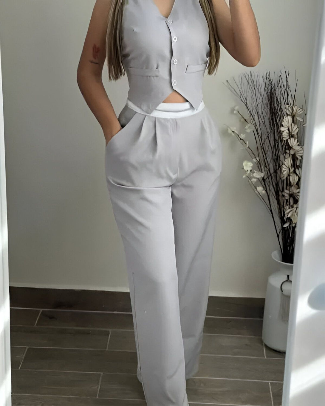 Une femme porte un tailleur gris avec un pantalon large et un gilet cintré. Elle a les cheveux longs et blonds et porte un maquillage léger. Elle est debout dans une pièce avec un mur blanc et un sol en carrelage.