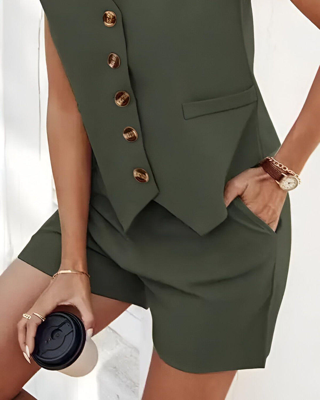 Une femme porte un gilet vert kaki et un short assorti. Elle a une main dans la poche du short et l'autre tient une tasse à café.