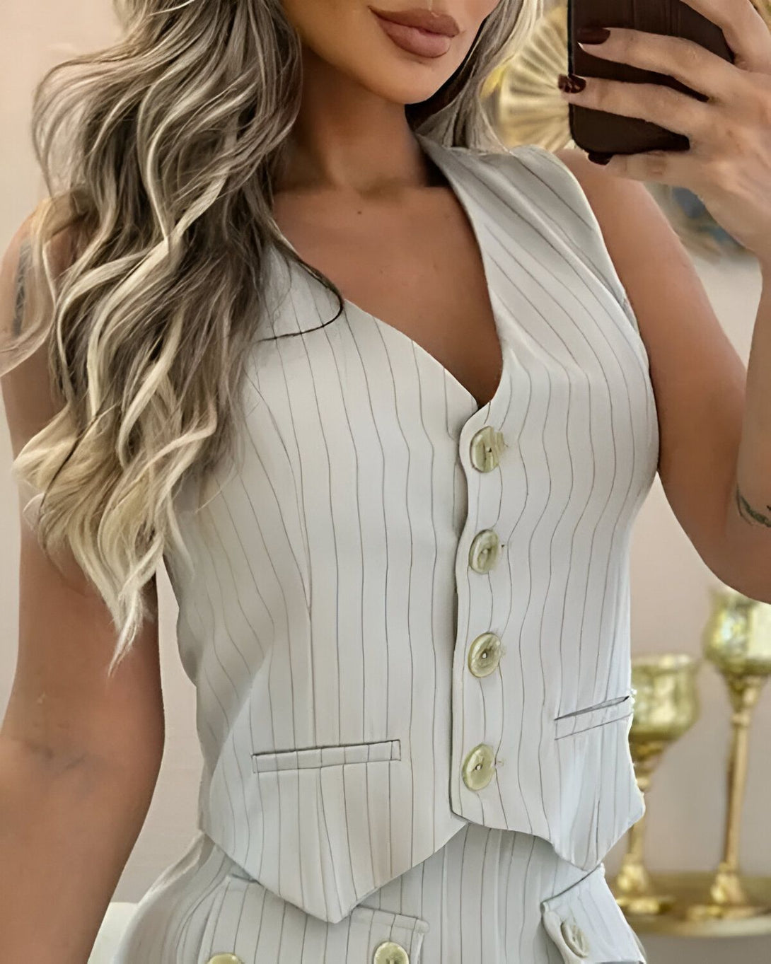 Une femme porte un gilet beige et un pantalon assorti. Le gilet est cintré et possède des boutons sur le devant. Le pantalon est taille haute et possède des poches sur les côtés. La femme est blonde et a les cheveux longs. Elle est maquillée et porte un rouge à lèvres nude. Elle est debout et tient son téléphone portable dans sa main droite.