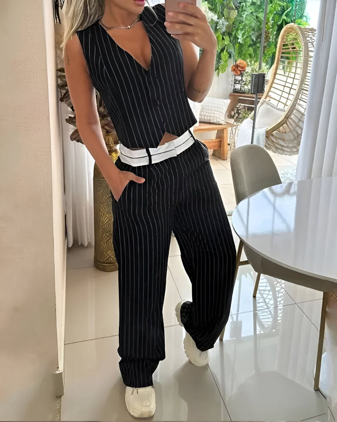 Une femme porte un gilet noir et blanc à rayures verticales, un pantalon noir et blanc à rayures verticales, et des baskets blanches. Elle a une main dans la poche de son pantalon et l'autre tient un téléphone portable.